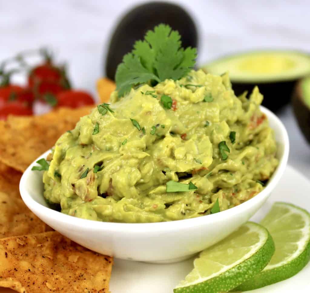 guacamole in white bowl with lime slices and tortilla chips on side
