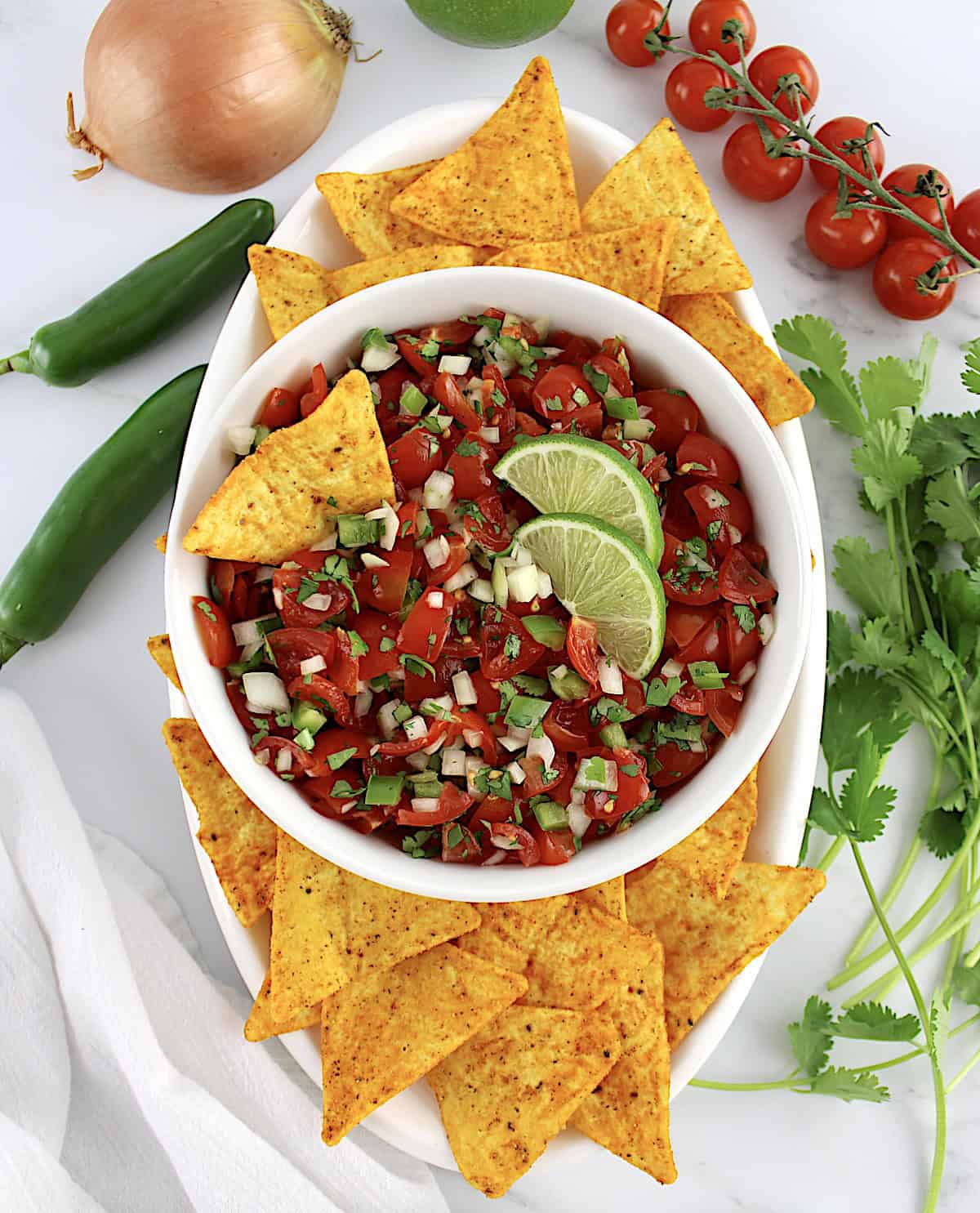 overhead view of Pico de Gallo in white bowl with tortilla chip and 2 lime slices with more chips around the bowl