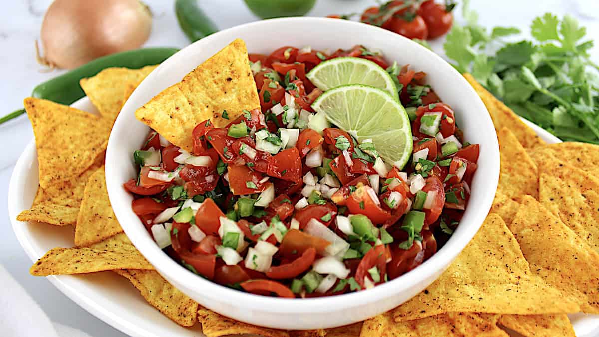 Pico de Gallo in white bowl with tortilla chip and 2 lime slices with more chips around the bowl